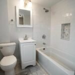 Small well lit white tile bathroom with toilet, sink and bathtub.